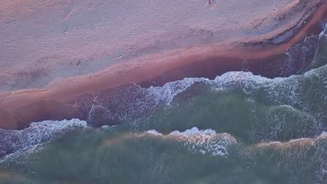 aerial birdseye view of the coastline of a sandy beach with gentle waves hitting the yellow sand, baltic sea in liepaja, latvia, golden hour light, wide angle point of view drone shot