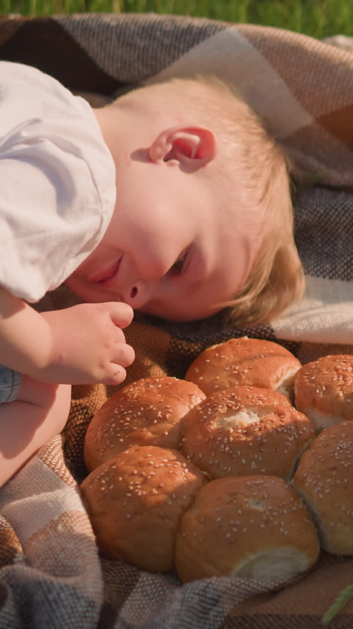 Two young boys wearing white shirts are lying on a checkered scarf in a grassy field, enjoying pieces of freshly baked bread.The peaceful outdoor setting highlights a simple serene moment of childhood