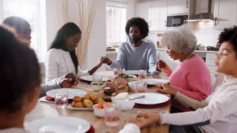 Multi-generation-mixed-race-family-holding-hands-and-saying-grace-at-the-table-before-eating-their-Sunday-dinner,-selective-focus