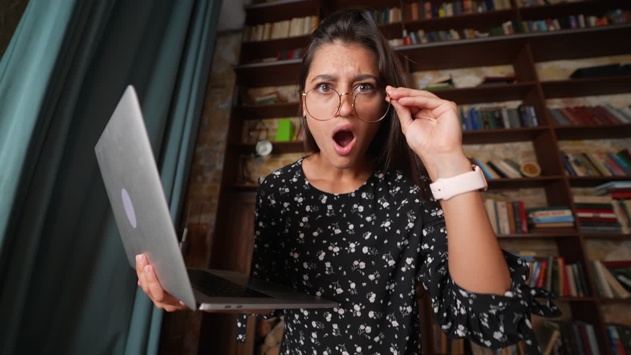 mujer con gafas, sosteniendo una computadora portátil, expresión de sorpresa en la biblioteca