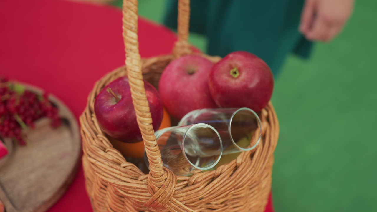 persona che sistema delle mele in un cesto di vimini su un tavolo rosso con occhiali da sole e un'arancia nelle vicinanze, le mani dispongono delicatamente la frutta su uno sfondo verde, atmosfera accogliente da picnic, dettaglio ravvicinato di uno stile di vita, estate fresca
