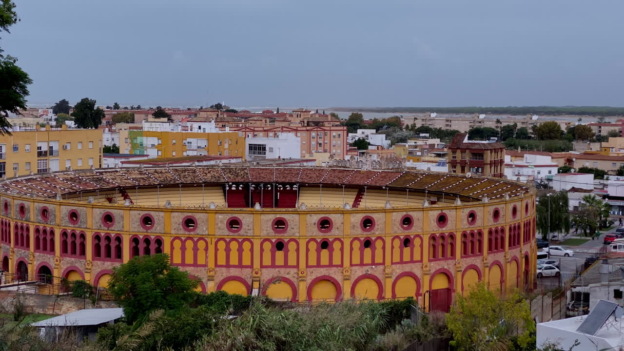 Una ladera abandonada con plantas secas enmarcando una plaza de toros tradicional roja y amarilla, y el pueblo circundante.