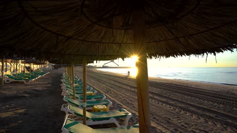 Gimal-POV-marbella-beach-at-sunrise-walking-inbetween-tropical-parasols-and-sunbeds,-silhouette-of-couple-walking-on-beach