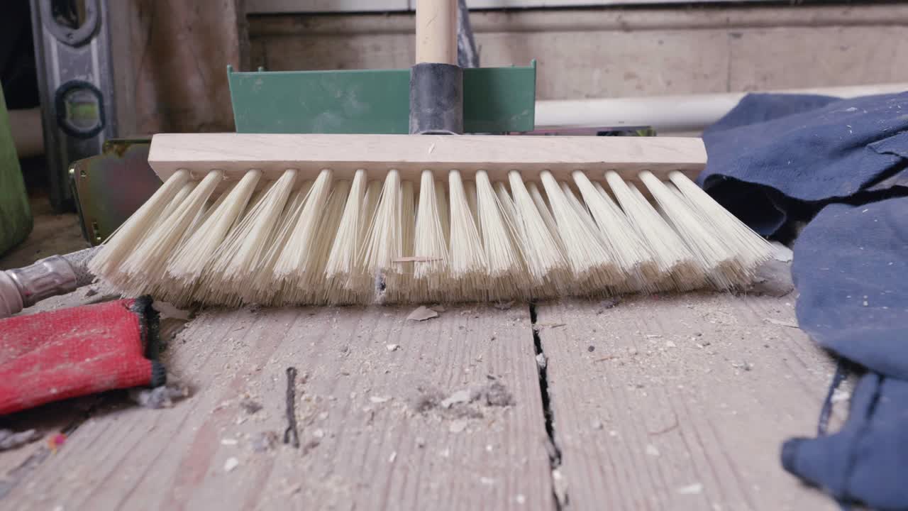 Bathroom renovation push in on broom head amidst dusty, bare wooden floorboards, working gloves, drop cloth and spirit level