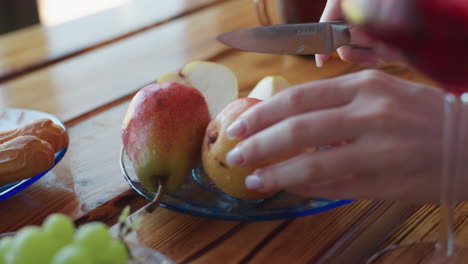close up of hand adjusting transparent plate holding fresh pears and sliced apple on wooden table, surrounded by grapes, pastries, and red drink in elegant glass, all under warm natural lighting