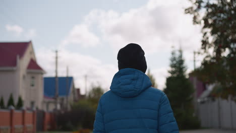 close up back view of person in blue jacket and black beanie strolling in residential area, with blurred houses and trees in background under cloudy skies