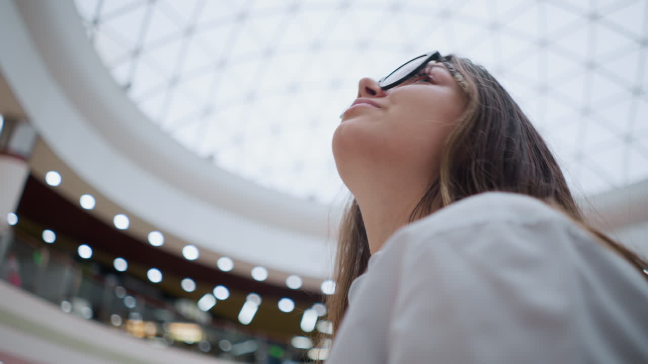 primer plano de una joven con gafas en una escalera mecánica en un centro comercial concurrido, en un estado de ánimo reflexivo mientras mira hacia la derecha, capturando su expresión contemplativa