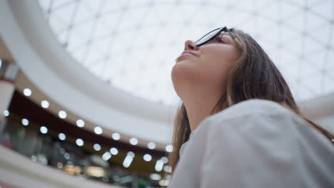 primer plano de una joven con gafas en una escalera mecánica en un centro comercial concurrido, en un estado de ánimo reflexivo mientras mira hacia la derecha, capturando su expresión contemplativa