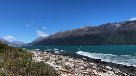 Two-kite-surfers-are-pulled-along-by-the-strong-wind-in-the-distance-in-front-of-a-beach,-waves,-and-mountain-range-in-Lake-Wakatipu-in-New-Zealand