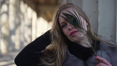 blond model woman posing for photoshoot with flashlight outdoors holding peacock feather covering face