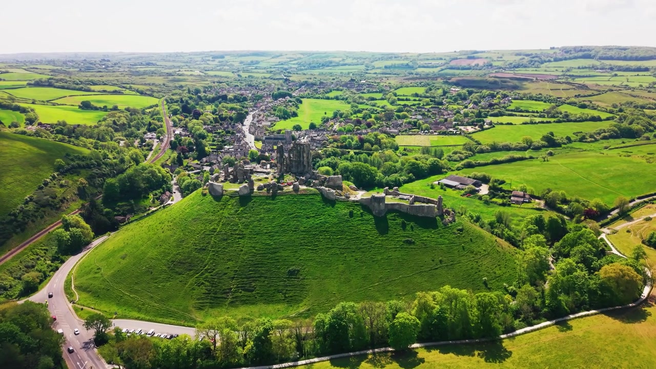 Nagranie z drona przedstawiające ruiny zamku Corfe stojące na zielonym wzgórzu nad otaczającą wioską i okolicą w Dorset w Anglii. Szerokie ujęcie przedstawia historyczny zamek, wiejski krajobraz, pola
