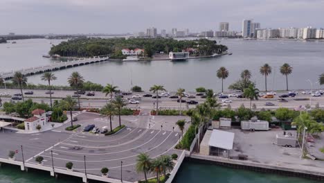 Vista-Cinematográfica-De-Una-Carretera-Con-Autos-En-Movimiento-Y-Tráfico-Frente-A-La-Isla-De-Palmeras-Y-El-Paisaje-Urbano-En-El-Fondo-Vista-Aérea-|-Toma-De-Drone-De-Una-Ciudad-Con-Una-Carretera-Y-Una-Isla-De-Palmeras-En-Florida