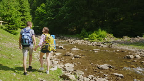 A-Young-Couple-Of-Tourists-Walking-Along-A-Mountain-River-Against-A-Background-Of-Green-Forest-Trave