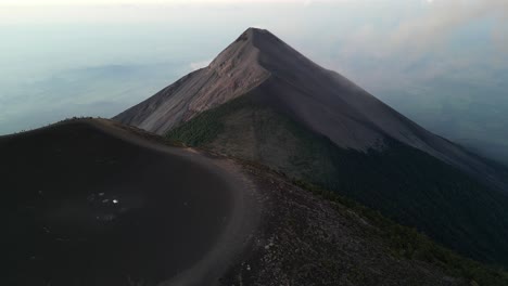 Drone-view-in-Guatemala-flying-over-a-volcano-crater-covered-in-ashes-at-sunrise-surrounded-by-green-mountains-and-clouds-over-towns