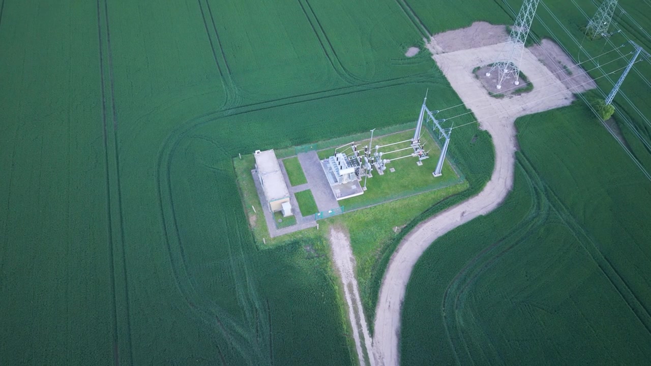Top-down drone shot of an electrical substation surrounded by green farmland in Germany, connected to renewable wind energy infrastructure and modern power distribution systems in UHD.