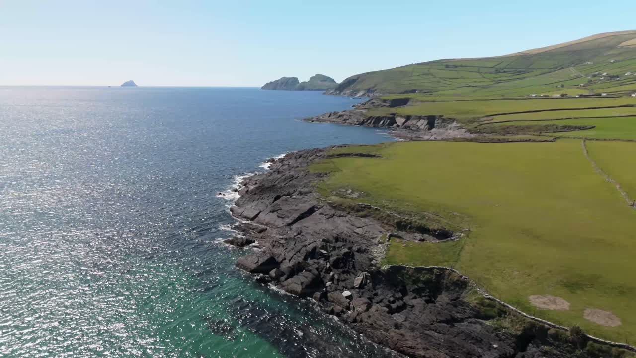 Aerial flight over the rugged coastline of Skellig Ring, Ireland. Cliffs drop into calm blue ocean with lush greenery and distant Skellig Island in view. Wild and majestic.