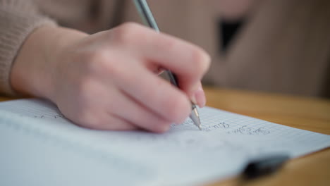 close-up of student taking notes in notebook with pen on woven cloth placed on wooden table, focused writing activity with soft lighting and natural wooden surface