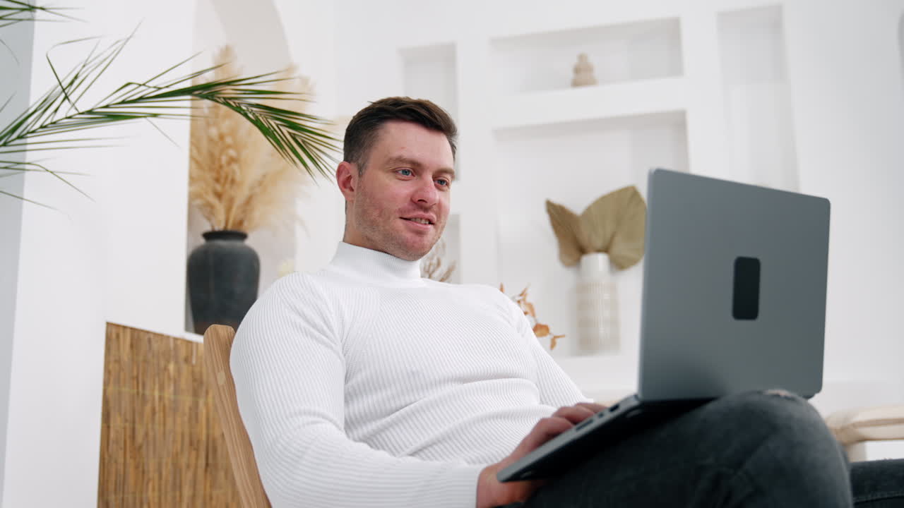 Positive male in white sweater sits holding the laptop. Freelancing man working from home. Low angle view.