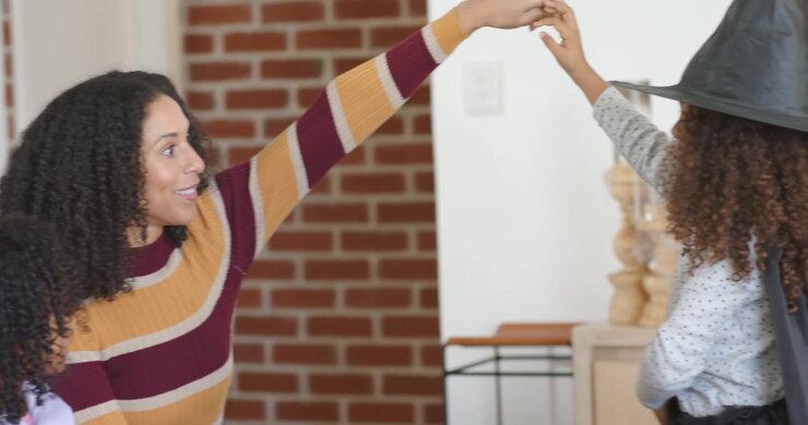 Mamma afroamericana che allunga la mano sinistra per guidare una ragazza con cappello da strega e mantello a casa, assicurandosi che vesta bene.