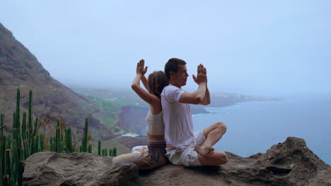 posicionados en la roca de una montaña, un hombre y una mujer meditan y practican yoga espalda con espalda, con la inmensidad del océano detrás de ellos