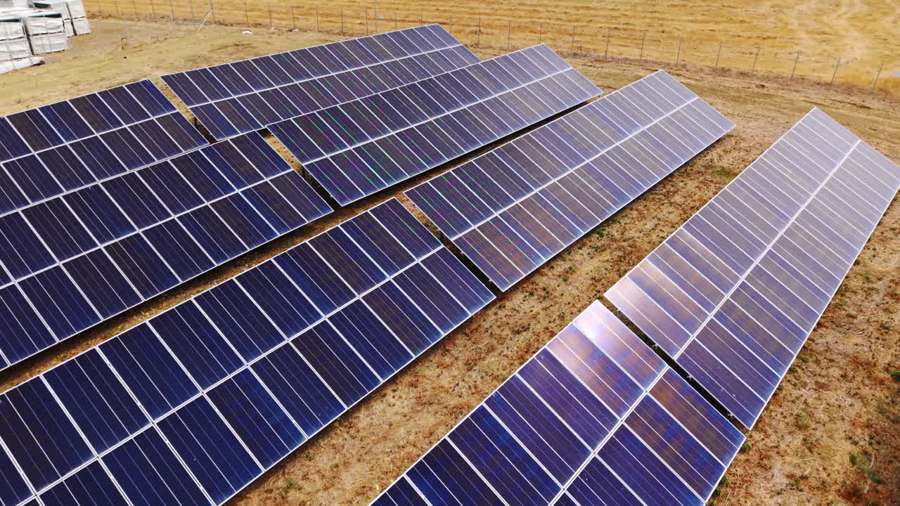 Drone flying very low over grouped photovoltaic solar panels, reflecting sunlight, with surrounding field fences visible in Sicily