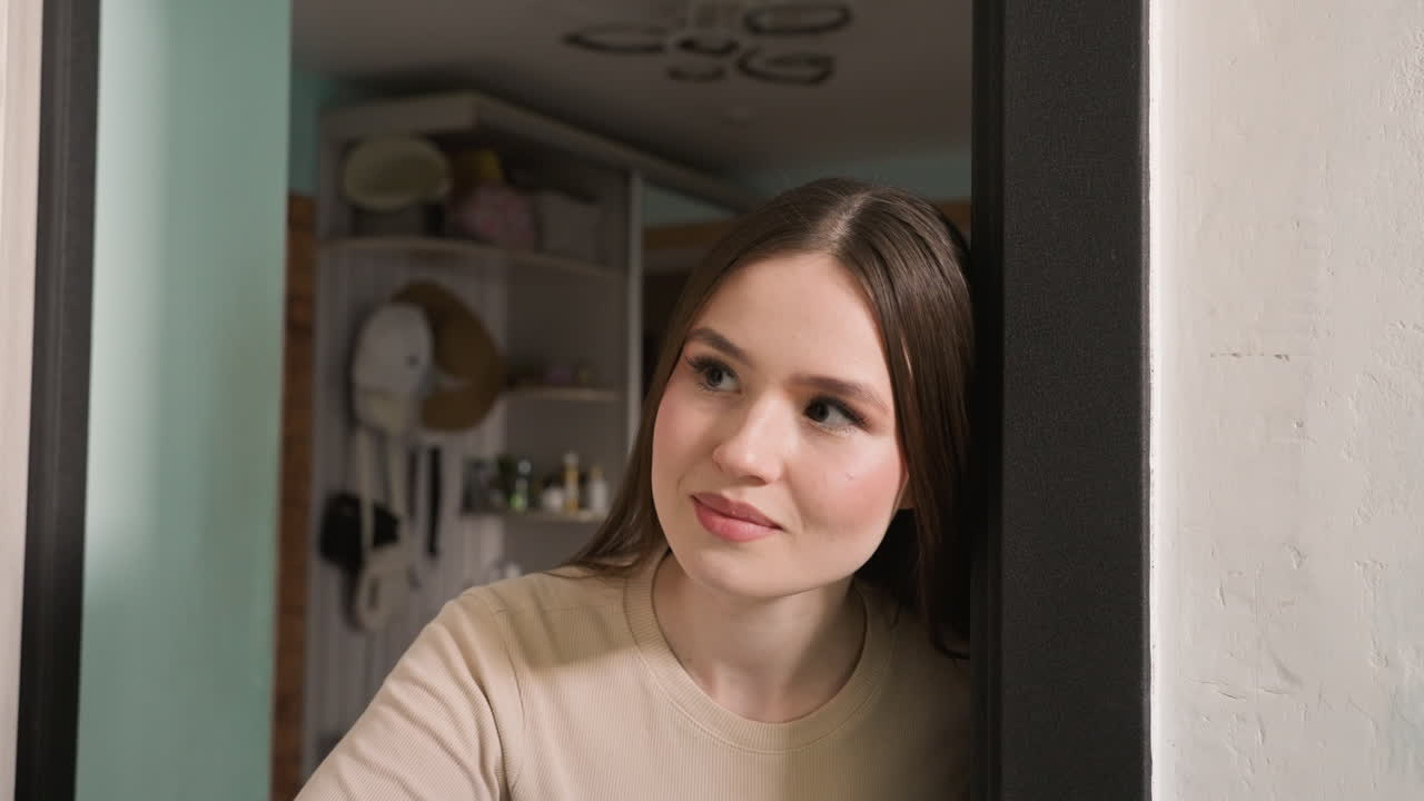 Giovane donna bianca che guarda attraverso la porta in un corridoio, sbirciando per controllare consegne e visitatori, luce naturale soffusa, maglione neutro, scaffali accoglienti della cucina visibili, espressione attenta, mattinata domestica