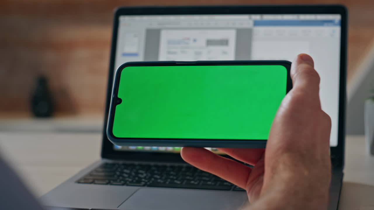 Primer plano del dedo de un hombre tocando un smartphone con pantalla verde en el escritorio de trabajo con laptop de la oficina.