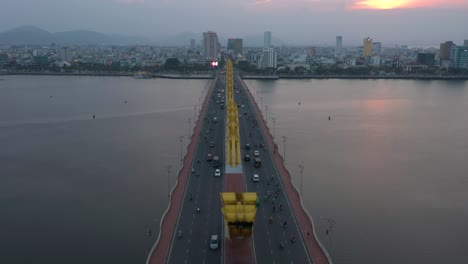 Drone-flying-backwards-over-Dragon-Bridge-Cau-Rong,-traffic-and-city-skyline-during-sunset-in-Danang,-Vietnam