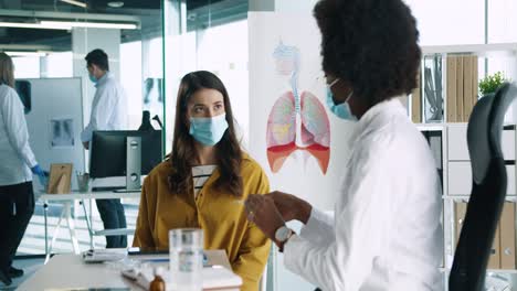 vista lateral de una doctora afroamericana con mascarilla médica sentada en el trabajo explicando a una paciente el tratamiento del coronavirus en consulta médica