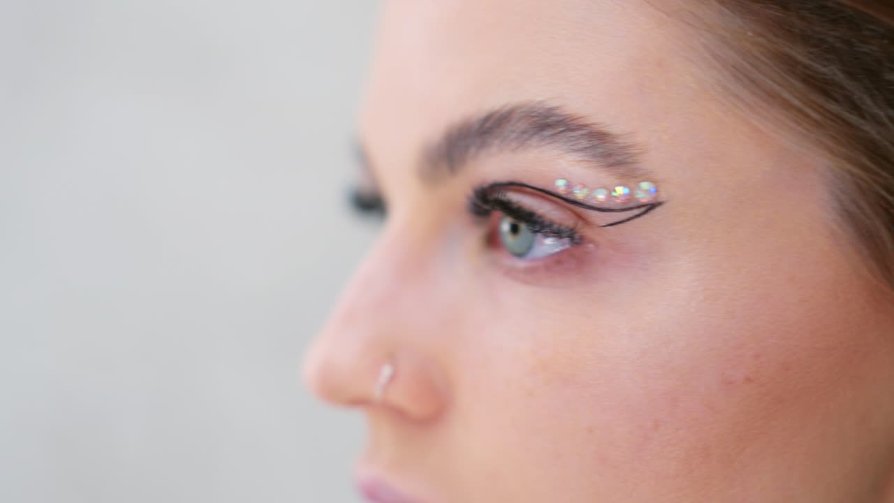 Close-up shots of an elaborate eye makeup application featuring graphic eyeliner and sparkling rhinestones