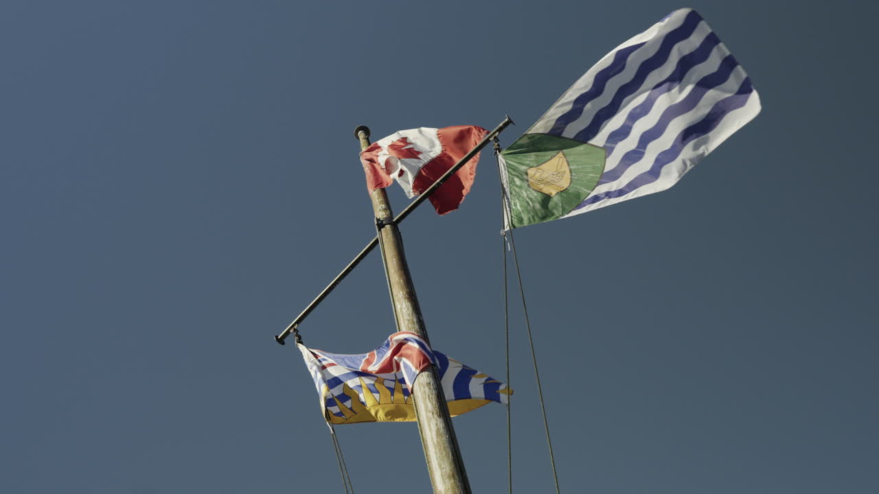 The flags of Canada, British Columbia and Vancouver on a mast waving in the wind, slow motion handheld, blue sky