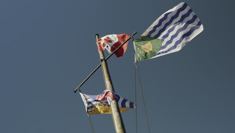 The-flags-of-Canada,-British-Columbia-and-Vancouver-on-a-mast-waving-in-the-wind,-slow-motion-handheld,-blue-sky