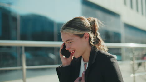 cheerful woman in formal attire smiling brightly during phone call while heading to office, hand placed on chest, enjoying joyful conversation, sunlight reflecting off modern glass building
