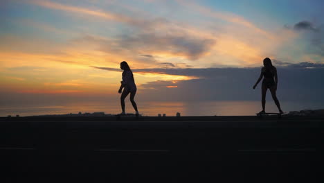In-a-slow-motion-scene,-two-girls-skateboard-on-a-road-at-sunset,-with-mountains-and-a-picturesque-sky-forming-the-backdrop.-Both-are-dressed-in-shorts