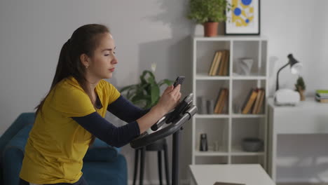 mujer morena está navegando por internet con su teléfono inteligente durante el entrenamiento con bicicleta estacionaria en casa adicción a la red social de los jóvenes modernos retrato medio en el interior