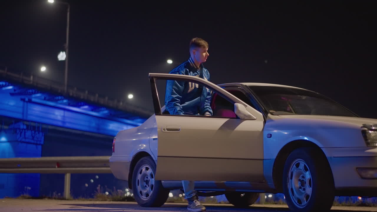 paso subterráneo nocturno, conductor caucásico se acerca al coche, mensajero solitario inspecciona el cupé, corredor callejero comprueba el retrovisor, mecánico de turno abre la puerta, tenso encuentro bajo las luces azules de neón del puente, asfalto