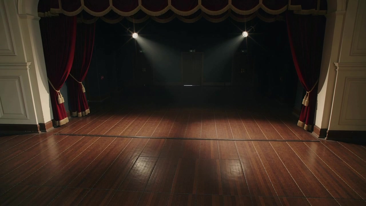 Luci dall'alto che si alzano e si allargano, rivelando un pavimento lucido incorniciato da tende rosse, durante i preparativi sul palco