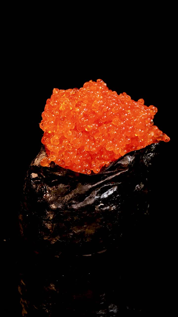 Sushi estilo Gunkan coberto com ovas de salmão laranja vibrante gira suavemente sob iluminação de estúdio dramática, em um fundo preto contínuo para um efeito visual impressionante.