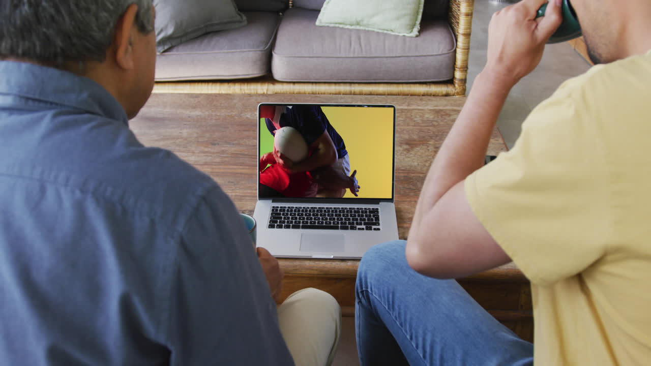 Padre e figlio birazziali che guardano un laptop con diversi giocatori di rugby maschi che giocano sullo schermo