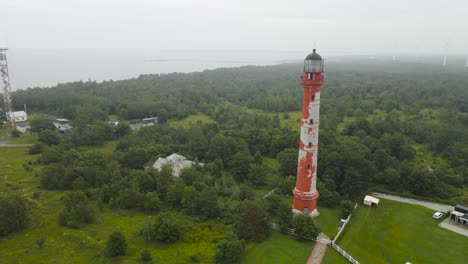aerial drone footage orbiting a historic red and white colored lighthouse in pakri paldiski peninsula during foggy and misty day while large white wind turbines and ocean sea water is in the back