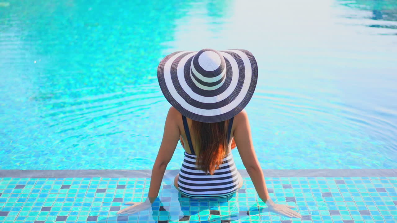 A beautiful young woman sits on the step of a large resort swimming pool, with her back to the camera