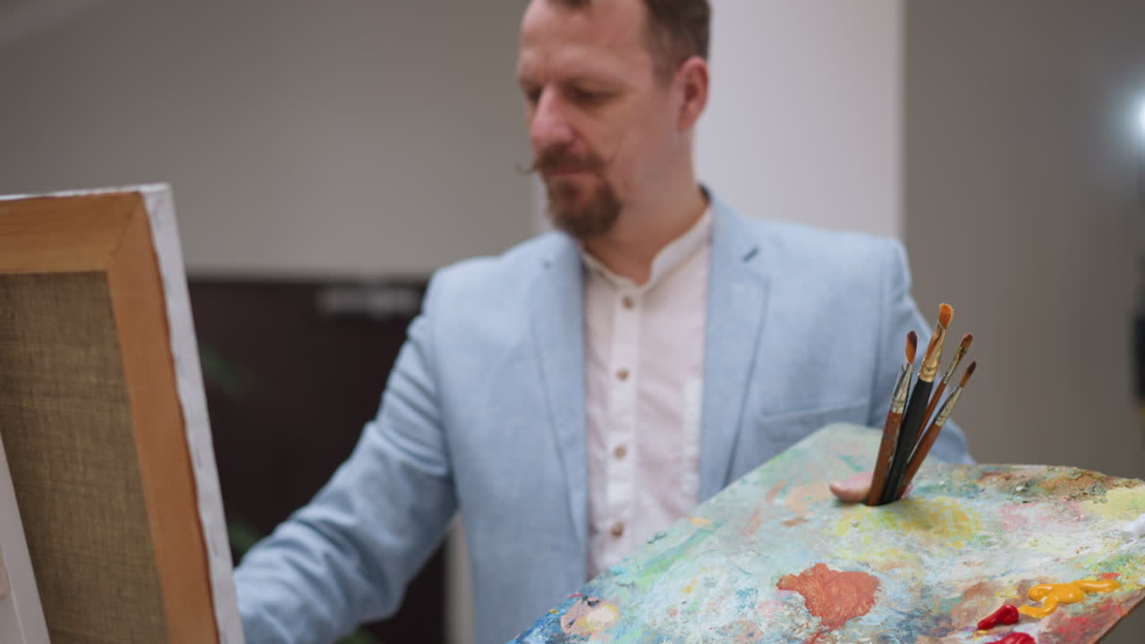 el artista dibuja sosteniendo una paleta rectangular en primer plano en el estudio. el creador de obras de arte con barba y bigote dibuja en el lienzo del caballete. el pintor elegante trabajando en la tienda