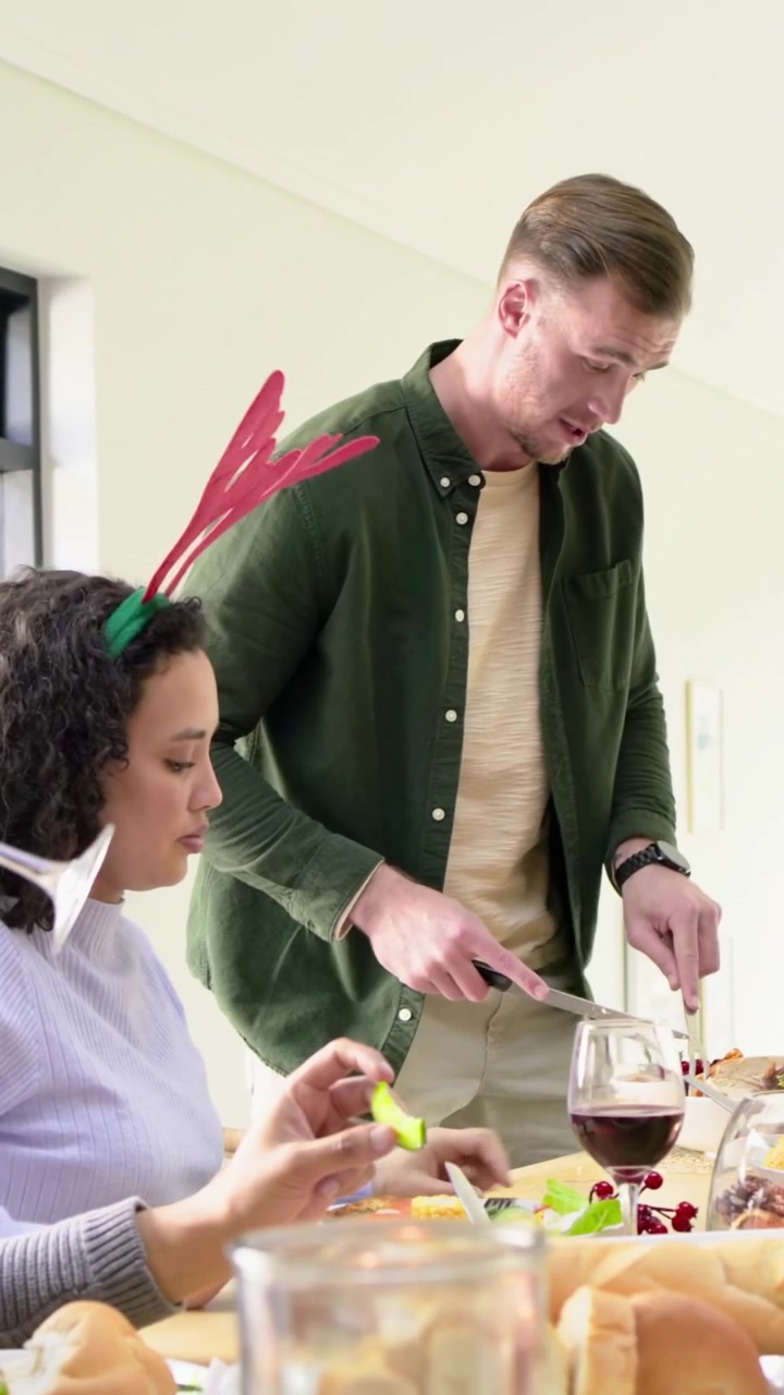 Vertical video: Reaching woman with antlers urging man carving roast with knife at table for feast