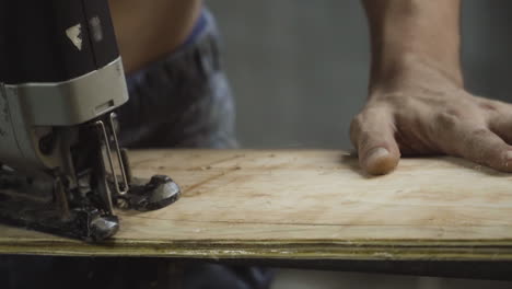 close up shot of a shirtless worker cutting a wooden ply board with a jigsaw machine and making a skateboard