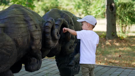 beautiful baby boy wearing white t-shirt and cap stands at the wooden sculpture. curious kid touches the figures in the park.