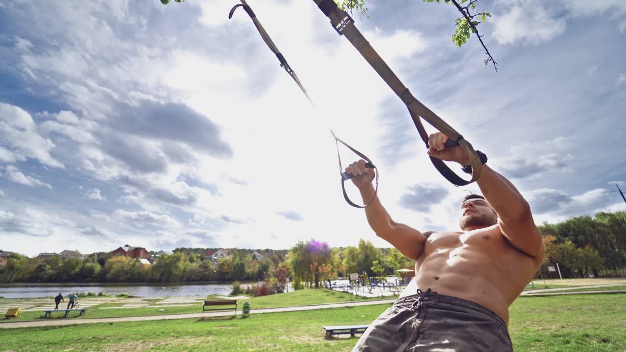Man is engaged in trx exercises under the sky. Muscular sportsman training his body with trx straps on beautiful nature background.
