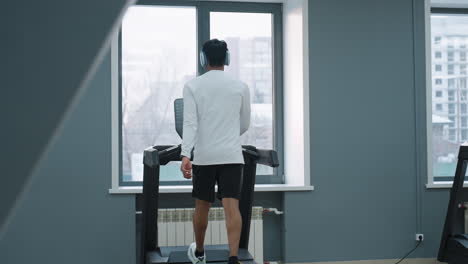 man in white shirt and black shorts walking on treadmill in bright gym with wall clock showing time and city buildings visible through windows for focused indoor cardio workout session