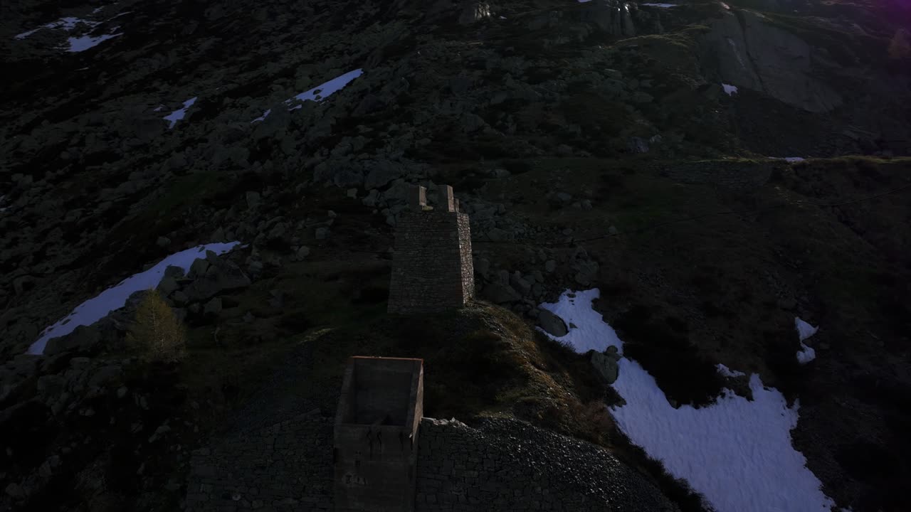 Moody aerial drone shot circling a historic stone tower ruin on a dark, rocky mountainside with patches of snow. Montespluga, Italian Alps, Lombardy, Italy