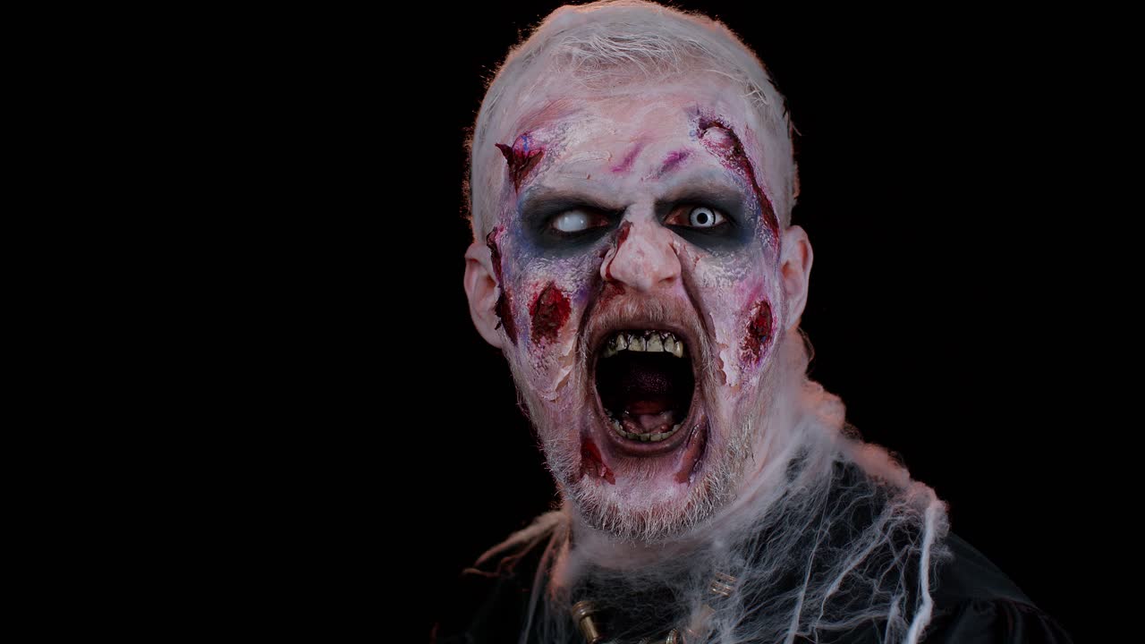 Zombie man with makeup with fake wounds scars and white contact lenses looking at camera, screaming