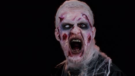 hombre zombi con maquillaje con heridas falsas cicatrices y lentes de contacto blancas mirando a la cámara, gritando